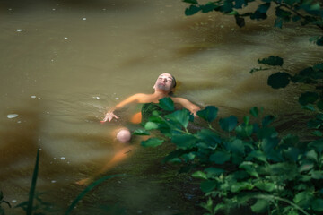 A young beautiful dark-haired girl swims in a pond in the shade of trees.