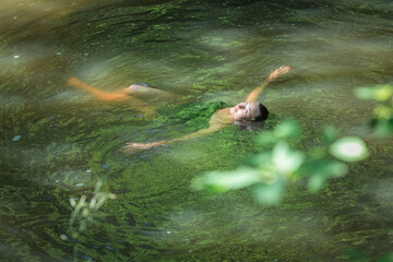 A young beautiful dark-haired girl swims in a pond in the shade of trees.