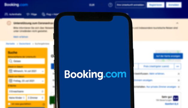 Booking.com logo is displayed on a modern smartphone, in the background the website of the online portal for booking hotels, apartments, flights, rental cars, vacations