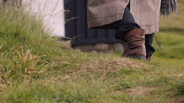 Mans Feet in Leather Boots Walks on Grass Towards Little Black House