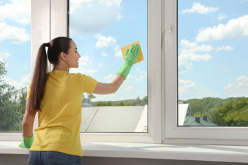 Happy young woman cleaning window glass with sponge cloth indoors - Powered by Adobe