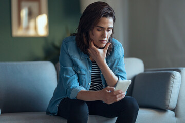 Shot of depressed woman using smartphone while sitting on the coach at home