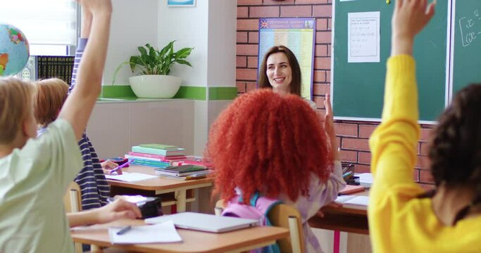 Rear on kids in school rising hands while answering to eacher in classroom. Back view. Children in class. Interesting lesson with young smiled lady teaching pupils. Indoor. Schooling concept.