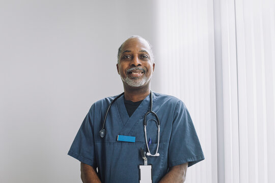 Portrait of smiling male doctor wearing stethoscope standing against white wall in medical clinic