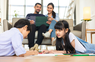 Happy Asian family relaxing on sofa while kid drawing on floor. Little boy girl having fun, friendship between siblings, family leisure time in living room at home.