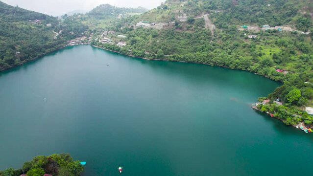 panning aerial shot over naukuchiatal lake bhimtal with it's blue green water with boats moving across and tree covered houses buildings on the side showing this tourist paradise in uttarakhand