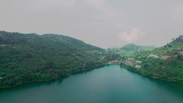 panning aerial shot over naukuchiatal lake bhimtal with it's blue green water with boats moving across and tree covered houses buildings on the side showing this tourist paradise in uttarakhand