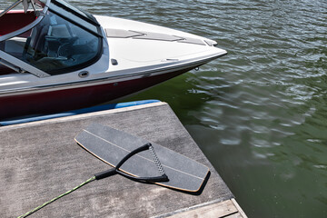 Wakeskate board with rope and handle lying next to a ski boat, wakeboard boat which is docked at the pier. A calm and empty lake in the background. Adventure, leisure, action sports scene.  - Powered by Adobe