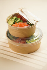 Trendy wholesome dish poke or buddha bowl - rice, wakame seaweed, tomatoes, cucumber, and red fish salmon - in a recycled round carton with sustainable fork, takeaway food concept on beige background