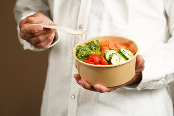 Man in white shirt holding Trendy dish poke bowl - rice, wakame seaweed, tomatoes, cucumber,...
