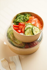 Trendy wholesome dish poke or buddha bowl - rice, wakame seaweed, tomatoes, cucumber, and red fish salmon - in a recycled round carton with sustainable fork, takeaway food concept on beige background