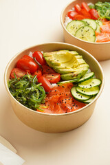 Trendy wholesome dish poke or buddha bowl - rice, wakame seaweed, tomatoes, cucumber, and red fish salmon - in a recycled round carton with sustainable fork, takeaway food concept on beige background