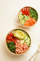 Trendy wholesome dish poke or buddha bowl - rice, wakame seaweed, tomatoes, cucumber, and red fish salmon - in a recycled round carton with sustainable fork, takeaway food concept on beige background