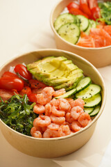 Trendy wholesome dish poke or buddha bowl - rice, wakame seaweed, tomatoes, cucumber, and red fish salmon - in a recycled round carton with sustainable fork, takeaway food concept on beige background