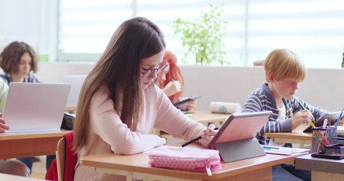 Caucasian schoolgirl in glasses sitting at desk in classroom, writing and using tablet device for study. Time at school. Smart girl learning at lesson and checking exercise in copybook with computer.