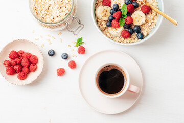 Coffee and homemade oatmeal with raspberries, blueberries, banana and chia seeds on white table. Healthy breakfast. Top view, flat lay
