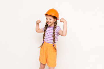 A little girl in a construction helmet and pigtails shows the strength of her biceps on a white isolated background. The profession of an engineer for a child.