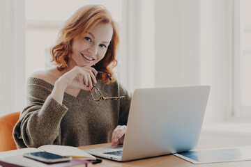 Pleased female scientist makes research on laptop computer, poses in coworking space, searches...