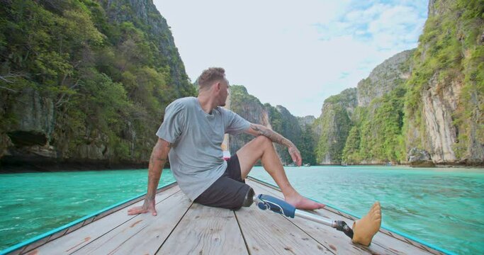 Young caucasian male with artificial bionic legs prosthesis sitting on the front of long tail boat seeing an amazing beautiful seascape during summer vacation travel in Phi Phi Islands, Thailand.
