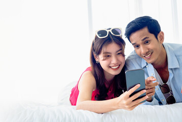 Happy Asian couple holds mobile phone. Young man and woman in casual feel happy with smiling and excited while having and looking at smartphone screen while lying on bed in white room with copy space.