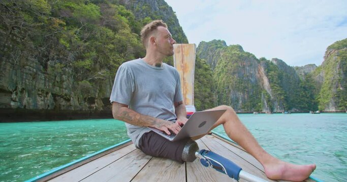 Disability freelancer caucasian man with artificial bionic legs prosthesis remote working anywhere on his laptop computer connected to office and relax by seeing beautiful view on summer vacation.