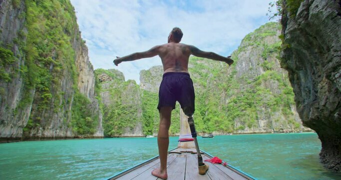Male with artificial bionic leg prosthesis standing on the front of long tail boat seeing an amazing beautiful seascape and raise his hand with a smile during summer vacation travel. Phi Phi, Thailand