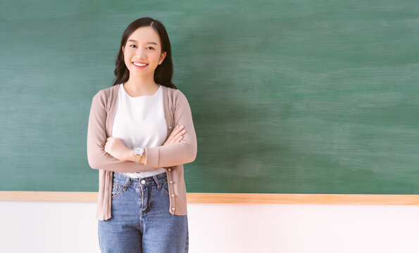Portrait of smiling beautiful Asian teacher standing with arms crossed looking at camera in front of a blank chalkboard in classroom. Back to school concept, with copy space