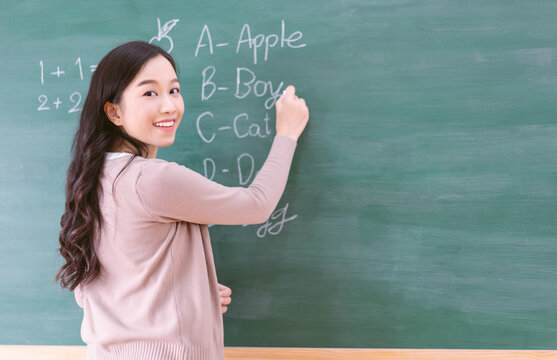 Portrait of smiling Asian teacher woman writing on chalkboard, teach english and math to students in classroom. Female teacher writing on blackboard wall and looking at camera. Back to school concept