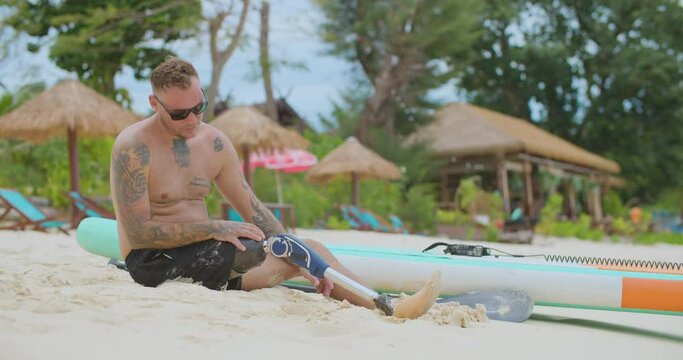 Tired surfer with bionic leg sitting on sandy beach. Front view of sportsman with artificial leg resting after surfing workout in ocean, looking at waves and relaxing. Sport, disability concept.