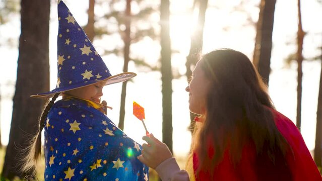 Childhood dreams, fantasies to become wizard. Child plays with his mother in costumes of wizard, playing in park on Halloween. Happy family. Child plays in mantle of magician, mother in red cloak.