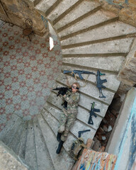 Caucasian woman in military uniform lies on the stairs of an abandoned building and holds a machine gun. View from above. 