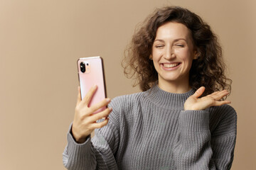 Friendly curly beautiful female in gray casual sweater doing video call with phone posing isolated...