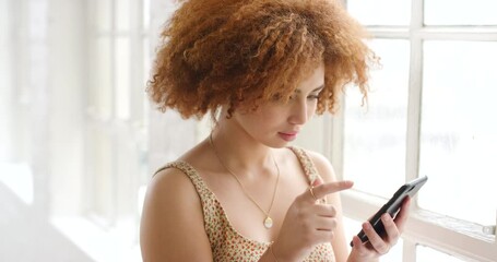 Young woman using a phone inside a bright room near a window. Trendy girl browsing online and checking social media on a mobile device. Single female swiping on a dating app looking for a partner