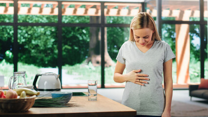 Beautiful Young Adult Woman with Blond Hair Wearing Gray V-Neck T-Shirt, Rubbing Her Stomach After Food. Healthy Female Drinking Daily Dose of Water at Spacious Home in Bright Living Room.