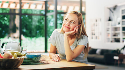 Portrait of Beautiful Young Adult Woman with Blond Hair Wearing Gray V-Neck T-Shirt, Leaning on a Table and Smiling Charmingly. Successful Woman Enjoying Leisure Time at Home in Bright Living Room.