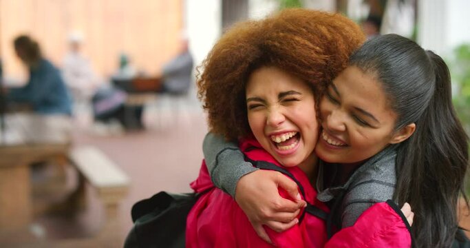 Cheerful best friends embracing and having fun while standing in a mall or at college. Two young multiethnic LQBTQ women laughing, hugging, and sharing a kiss on the cheek while meeting to catch up