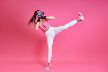 Young woman in virtual reality glasses kicking against pink background