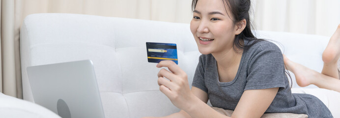 Fototapeta na wymiar Asian woman lounging on sofa and using laptop to shop online with credit card to register for payment or online transactions, Financial transactions and Internet security, Shopping with credit card.
