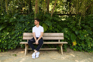 Handsome young man in white T-shirt and black trousers sitting on the wooden bench in the park...