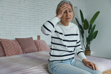 Frustrated senior woman touching forehead while sitting on the bed at home