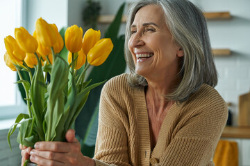Elegant senior woman holding a bunch of yellow tulips and smiling while relaxing at home - obrazy, fototapety, plakaty