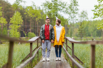 Fototapeta na wymiar Cheerful young couple wearing party hats standing on wooden path in countryside. Man looking at camera, woman grimacing. Birthday hike concept