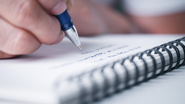 Close up hand writing paper on notebook with blue pen to note, memo, list action plan at office or home desk, selective focus. Concept of write on paper, letter, pen, notepad, diary