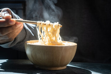 Hand uses chopsticks to pickup tasty noodles with steam and smoke in bowl on wooden background,...