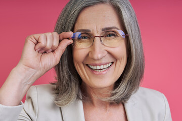 Obraz premium Happy senior woman in formalwear adjusting eyeglasses while standing against pink background