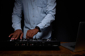 African American stylishly dressed man in DJ booth. On black background in the frame, only the body, close up. Professional DJ equipment and laptop on the table. On hand is a modern smart watch.