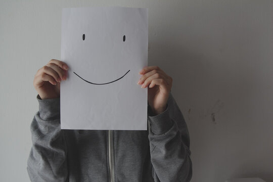 Woman holding a smiling face emoticon in front of face against a gray background, depression, mental health, masking feelings.