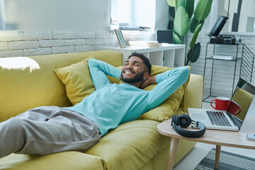 Happy African man holding hand behind head while relaxing on the couch at home