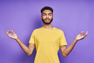 Young African man meditating with his eyes closed while standing against purple background - obrazy, fototapety, plakaty