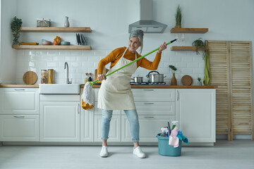 Playful senior woman carrying mop while standing at the domestic kitchen